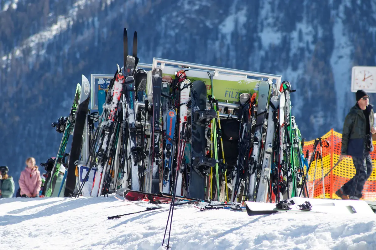 Stojak wypełniony nartami i snowboardami przy stoku, w tle ośnieżone góry i narciarze