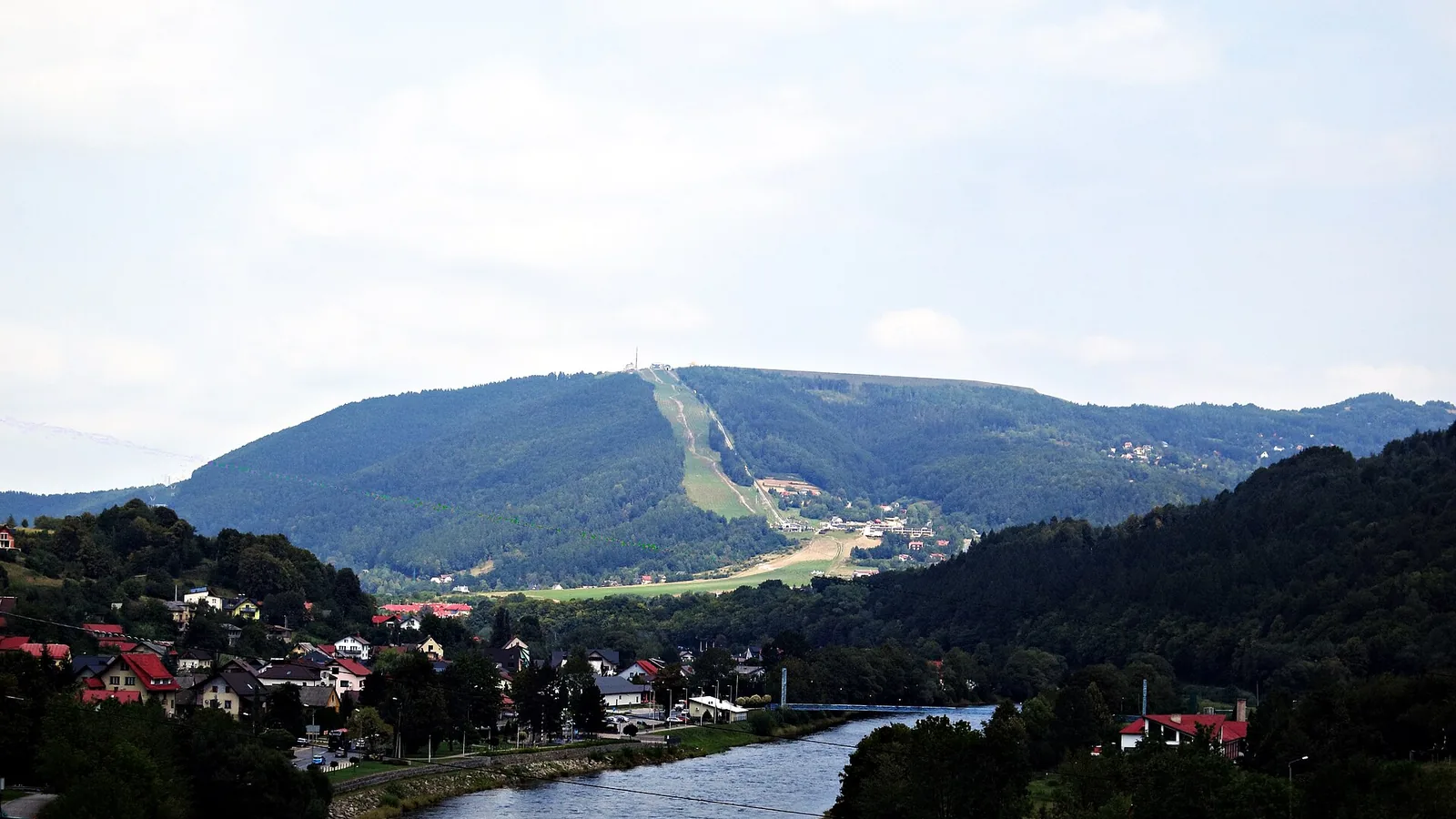 Góra Żar z wyraźną przecinką trasy zjazdowej nad zabudową i rzeką, widok od strony Tresnej
